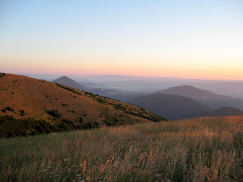Panoramic view of Monte Tezio Park at sunset, with meadows and mountain ridges stretching towards the horizon.