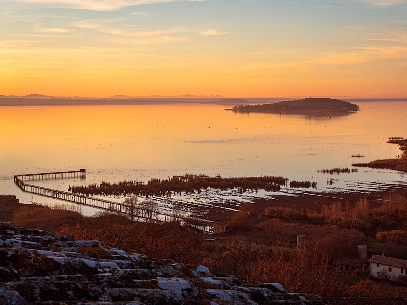 Panoramic view of Lake Trasimeno at sunset, with the water illuminated by the golden and orange hues of the sky and an island in the background. Credits: Azzurra Lucchetti