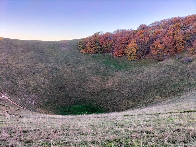 View of the Mortaro Grande on Mount Subasio, with its wide grassy depression surrounded by woodland in autumn shades of red, orange and yellow.