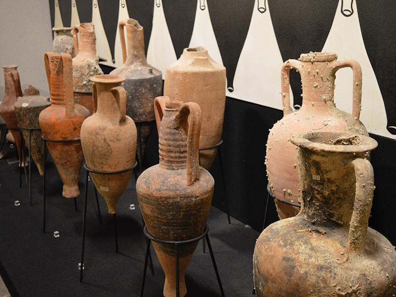 Amphorae and terracotta vessels on display, used in ancient times for the transport and storage of wine, against a black-and-white background.