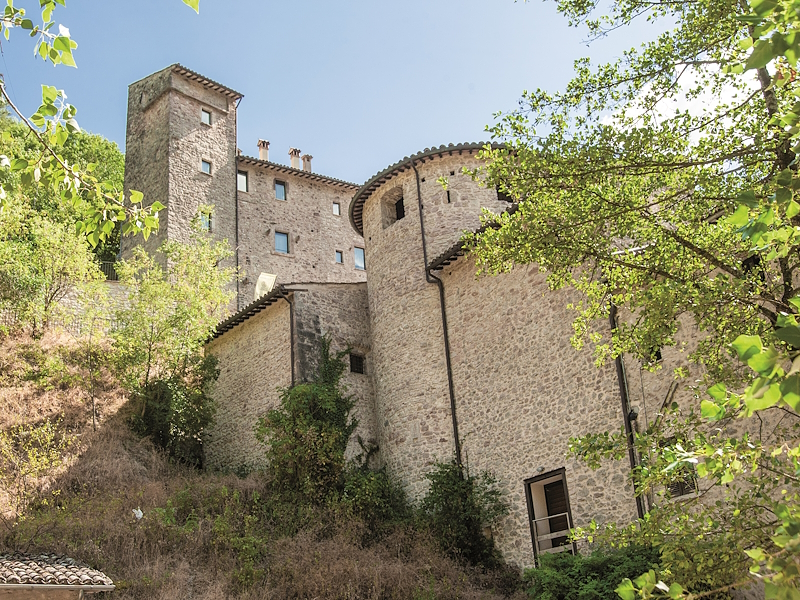 Steingebäude der Burg von Scheggino inmitten grüner Vegetation, mit Turm und befestigten Bauwerken am Hang.