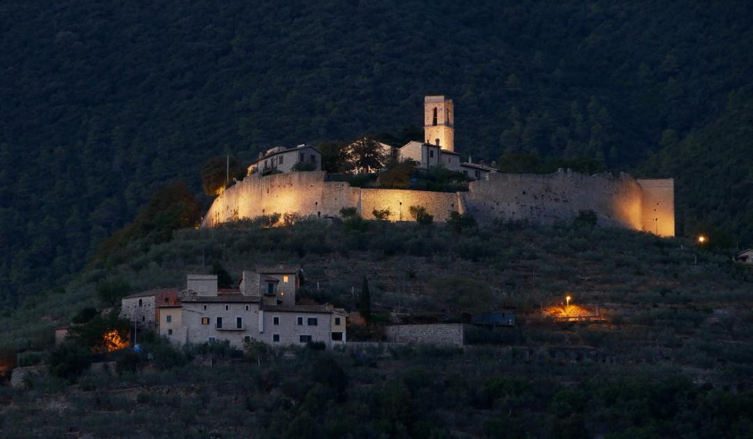 Il borgo fortificato di Campello Alto illuminato al crepuscolo, con mura e campanile che emergono dal pendio ricoperto di ulivi