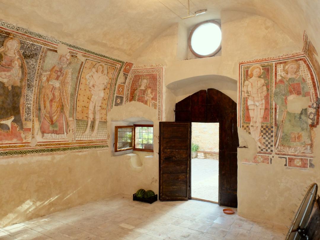 Interno affrescato con santi medievali e porta lignea aperta verso il cortile esterno