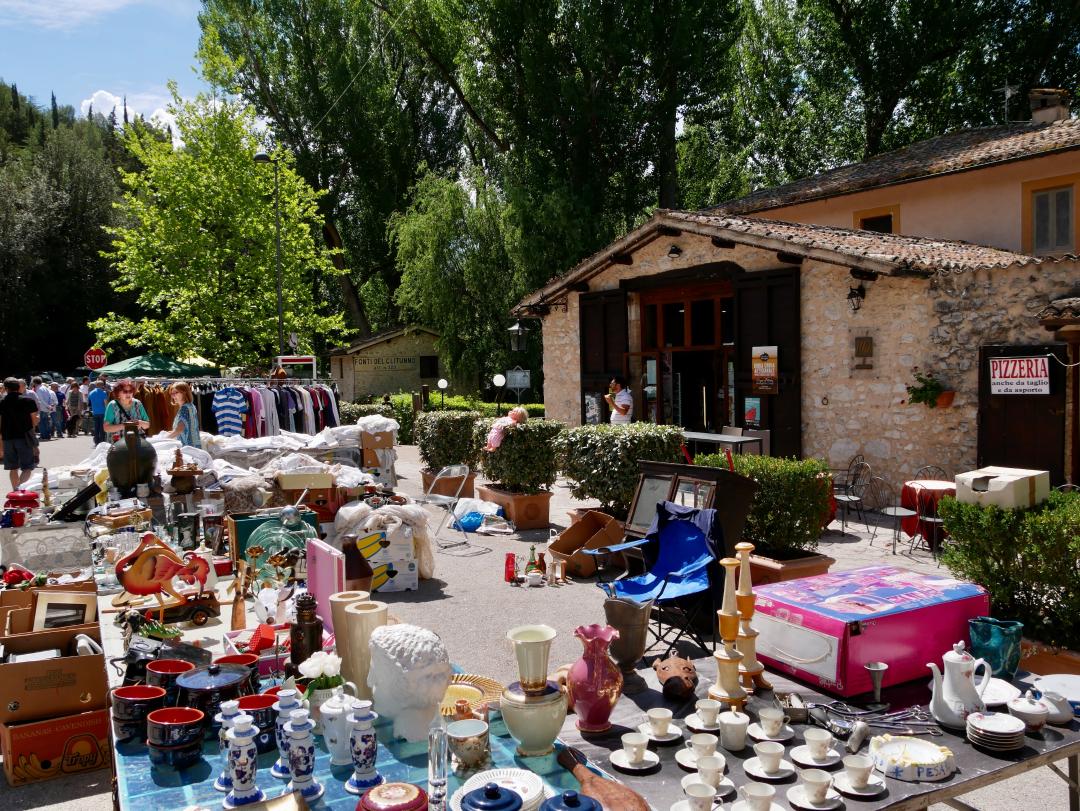 Stalls of an antique market with ceramics, vintage items and clothes displayed outdoors near the Fonti del Clitunno