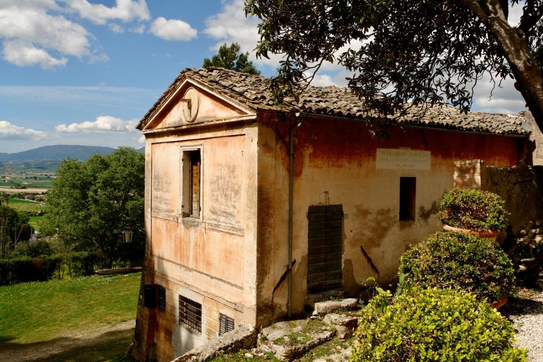 Old rustic building on a slope, with terracotta roof tiles and partially red-painted walls.