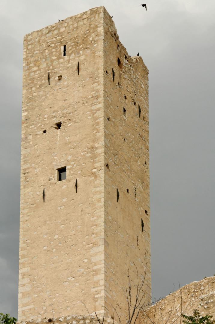 Tall medieval stone tower of Pissignano Castle photographed from below, with small arrow slits and swallows flying against a grey sky