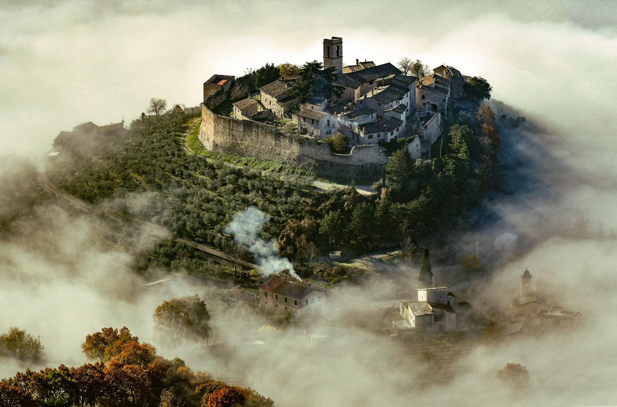 Campello Alto on a hill, surrounded by mist and olive groves, with medieval walls and stone houses emerging from the clouds