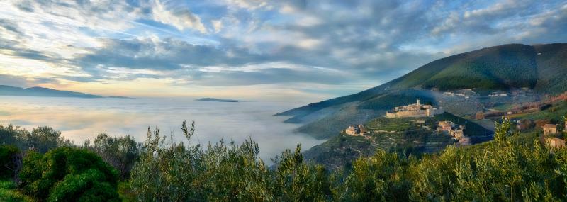 Village fortifié sur une colline entourée de brume, avec des maisons en pierre, des murs médiévaux et des oliveraies émergeant des nuages