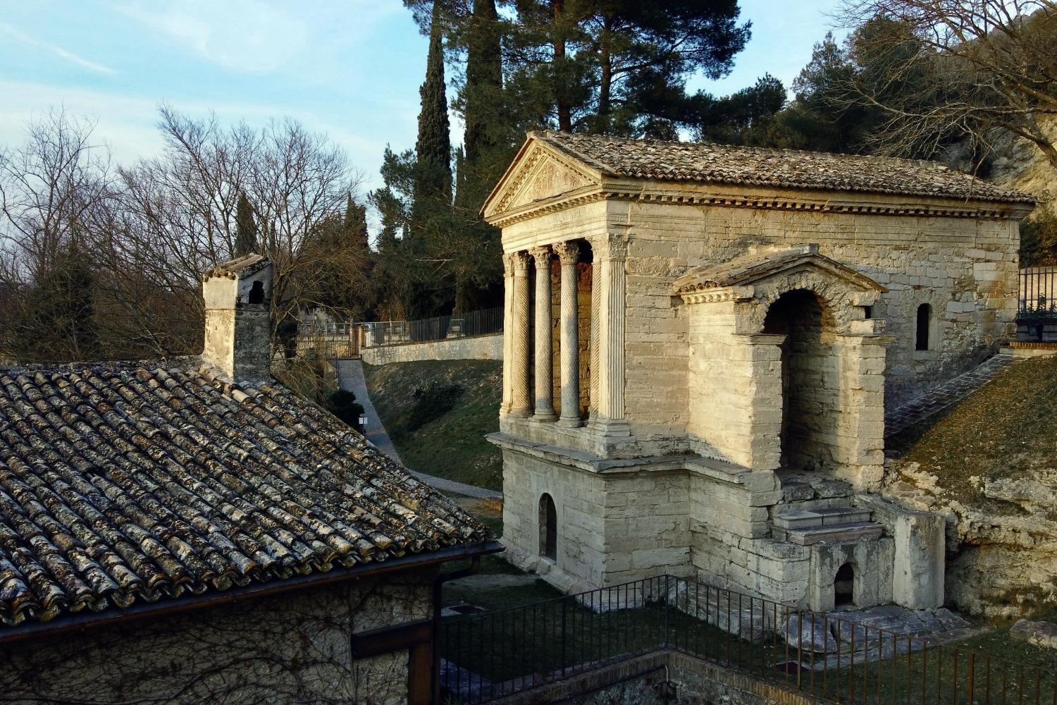 Side view of the Tempietto del Clitunno with Corinthian columns and a carved pediment, surrounded by trees and illuminated by the warm light of sunset