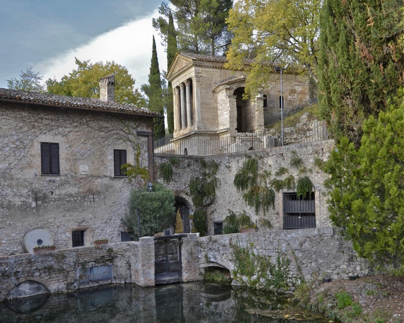 View of the Tempietto del Clitunno from the opposite riverbank, with a stone building in the foreground, surrounding vegetation, and reflections in the water