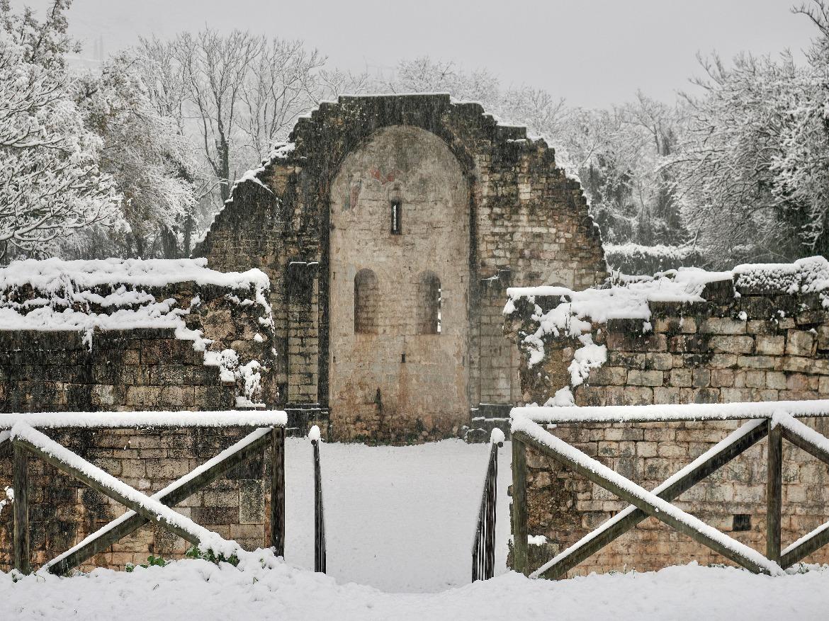 uined façade of the Church of San Cipriano covered in snow, framed by low walls and snow-covered trees.