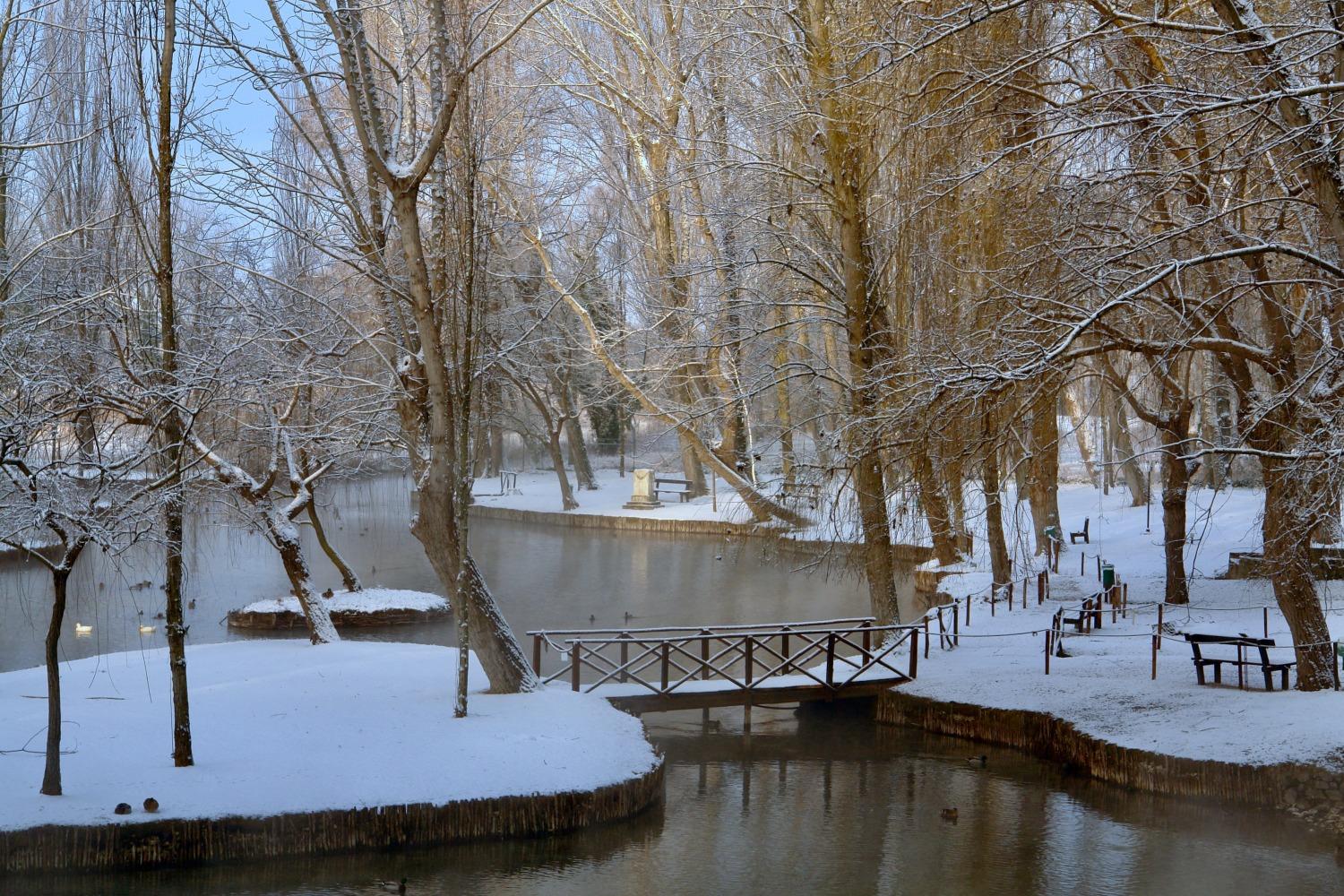 Snow-covered landscape with a wooden footbridge and frosted trees around the still water