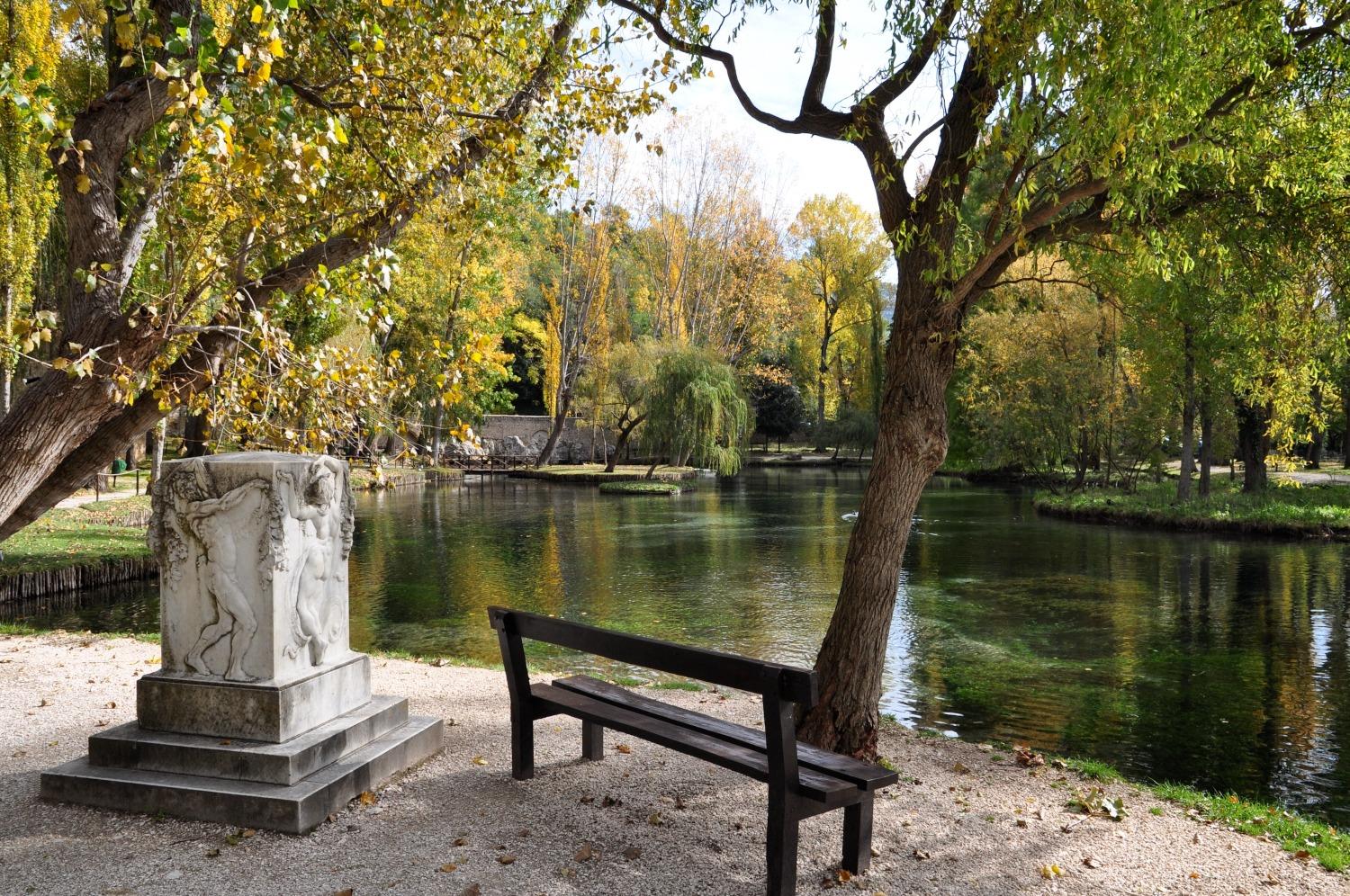Bench overlooking the lake beside a marble statue, surrounded by autumn trees and the clear waters of the Fonti del Clitunno