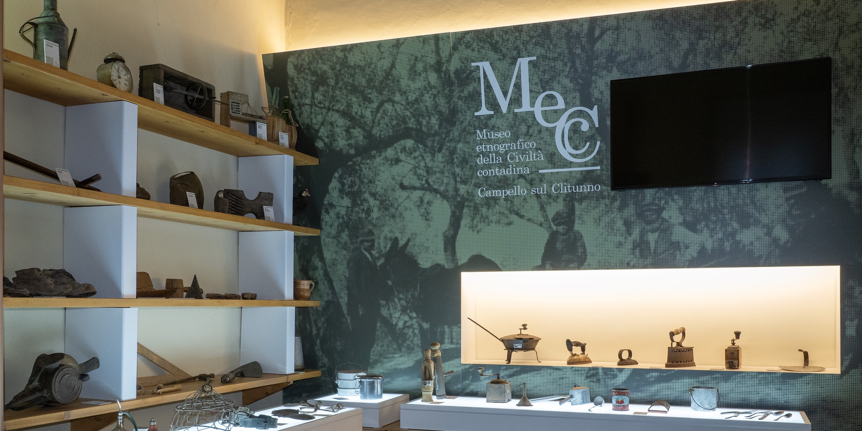 Exhibition of rural life objects with wooden shelves and a wall featuring the MeCC Museum logo