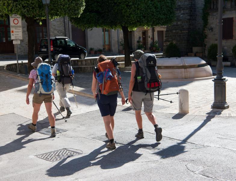 Group of pilgrims with backpacks and trekking poles crossing a historic square along the Via di Francesco.