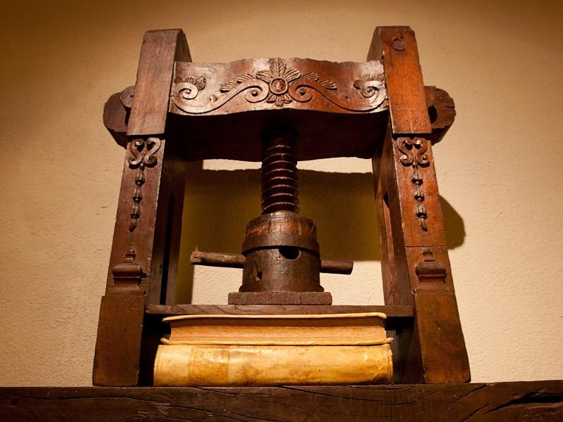 Antico torchio in legno con vite centrale e struttura decorata, esposto all’interno di uno spazio museale.