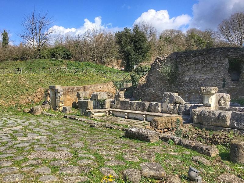 Resti archeologici di Ocriculum lungo l’antica Via Flaminia, con tratti di basolato romano, strutture murarie e frammenti lapidei immersi in un paesaggio verde.