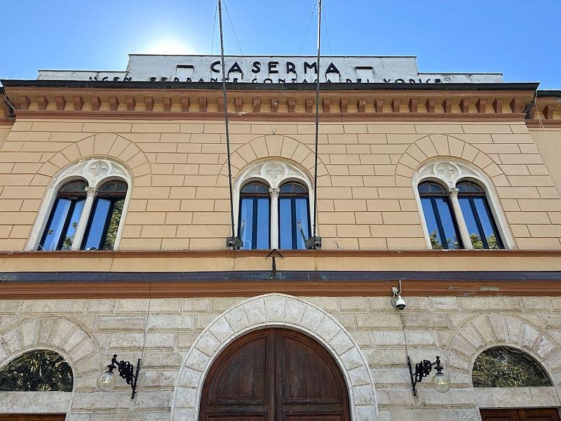 Façade of the Foligno barracks, with arched windows, a wooden entrance door and rooftop sign under a blue sky.