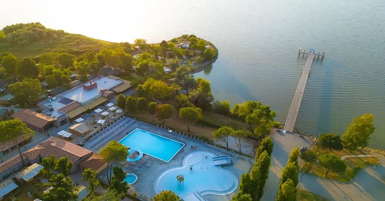 Vue aérienne d’un camping sur le lac Trasimène avec piscine, espaces de détente et un ponton s’étendant dans le lac, entouré de verdure.