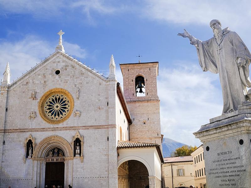 Statue of Saint Benedict in front of the basilica in Norcia, with stone facade and blue sky in the background