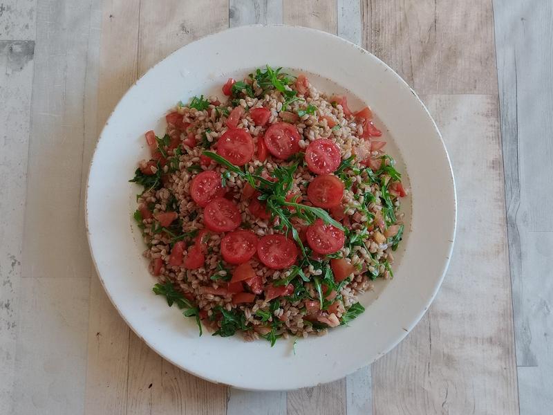 Plate of spelt salad with sliced cherry tomatoes and fresh arugula served on a light wooden table, top view