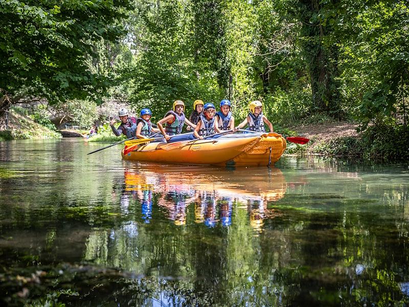 Groupe de personnes faisant du rafting sur un bateau pneumatique orange le long d’une rivière entourée de végétation, reflétées dans l’eau
